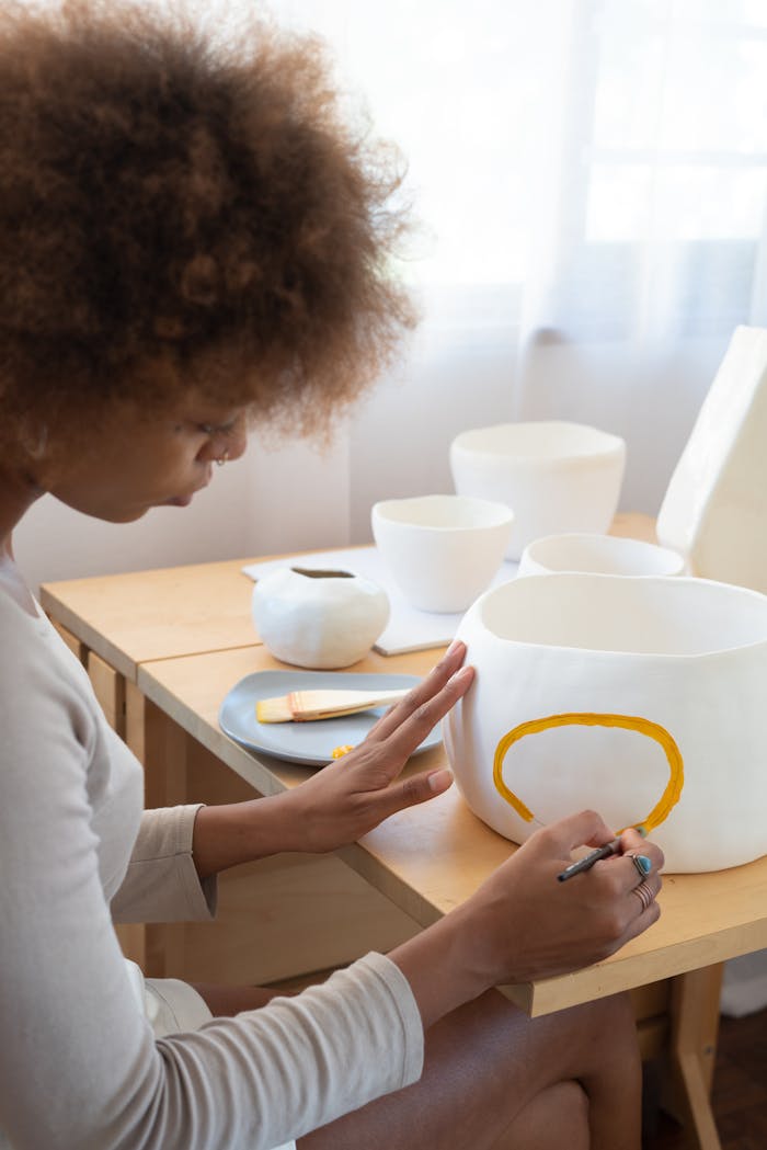 From above side view of talented African American female artisan sitting at table in creative workshop and paining picture on handmade ceramic bowl