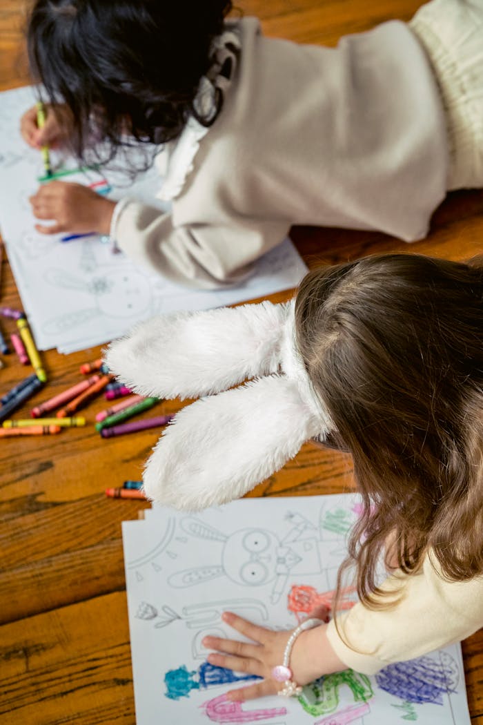 From above of crop faceless kids on wooden floor with wax pencil in hands coloring papers with Easter bunny and elements in light room